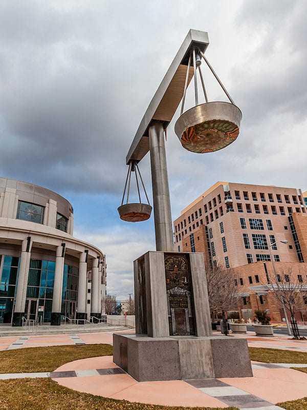 A large outdoor sculpture of a balance scale stands prominently in a plaza. The scale is made of metal, with two plates suspended on either side. It is positioned between a modern circular building and a rectangular office building under a cloudy sky.