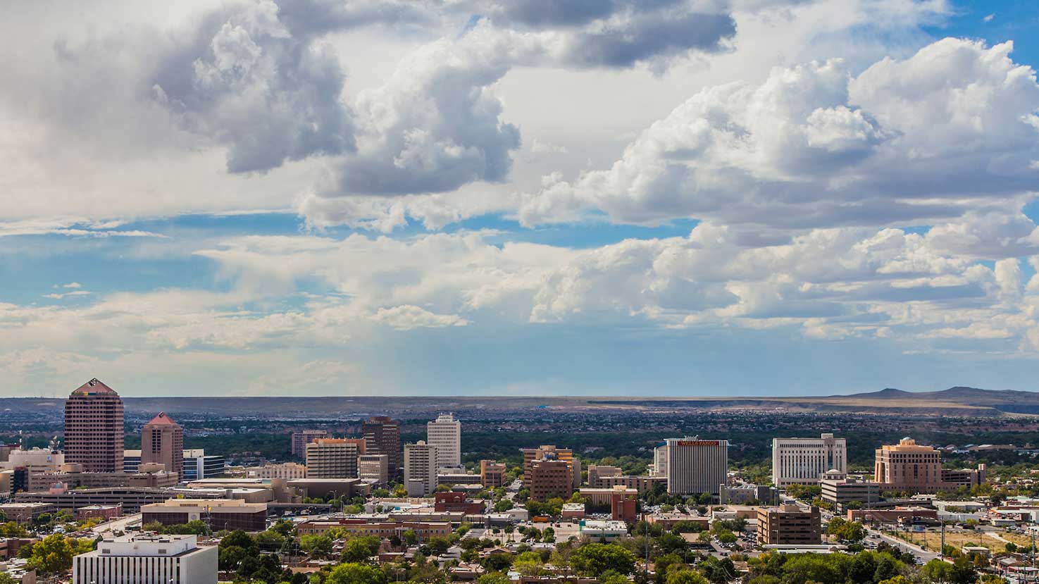 A cityscape view of Albuquerque, New Mexico, featuring a mix of modern buildings and greenery. The sky is partly cloudy with patches of blue visible. In the distance, the horizon stretches towards low hills.