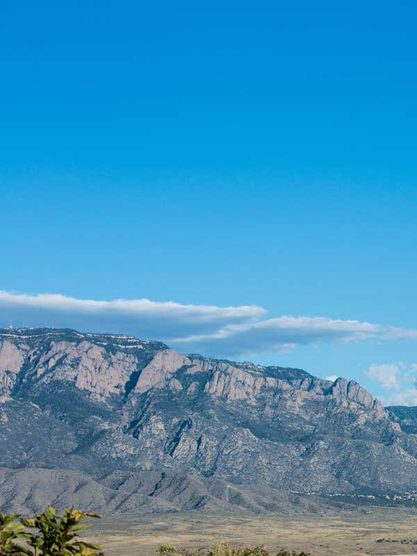 A scenic view of a mountain range under a clear blue sky. The rugged terrain features rocky slopes and a few scattered clouds in the distance. Vegetation is visible at the base of the mountains.