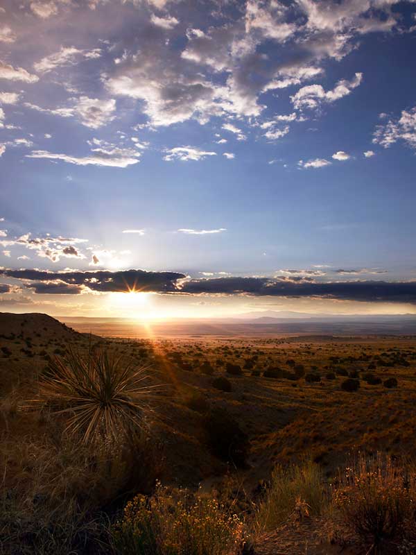 A landscape featuring a sunset over a vast desert. The sun is partially covered by clouds, casting a warm glow. The foreground has dry vegetation and sparse shrubs, with hills and a distant horizon visible under a partly cloudy sky.